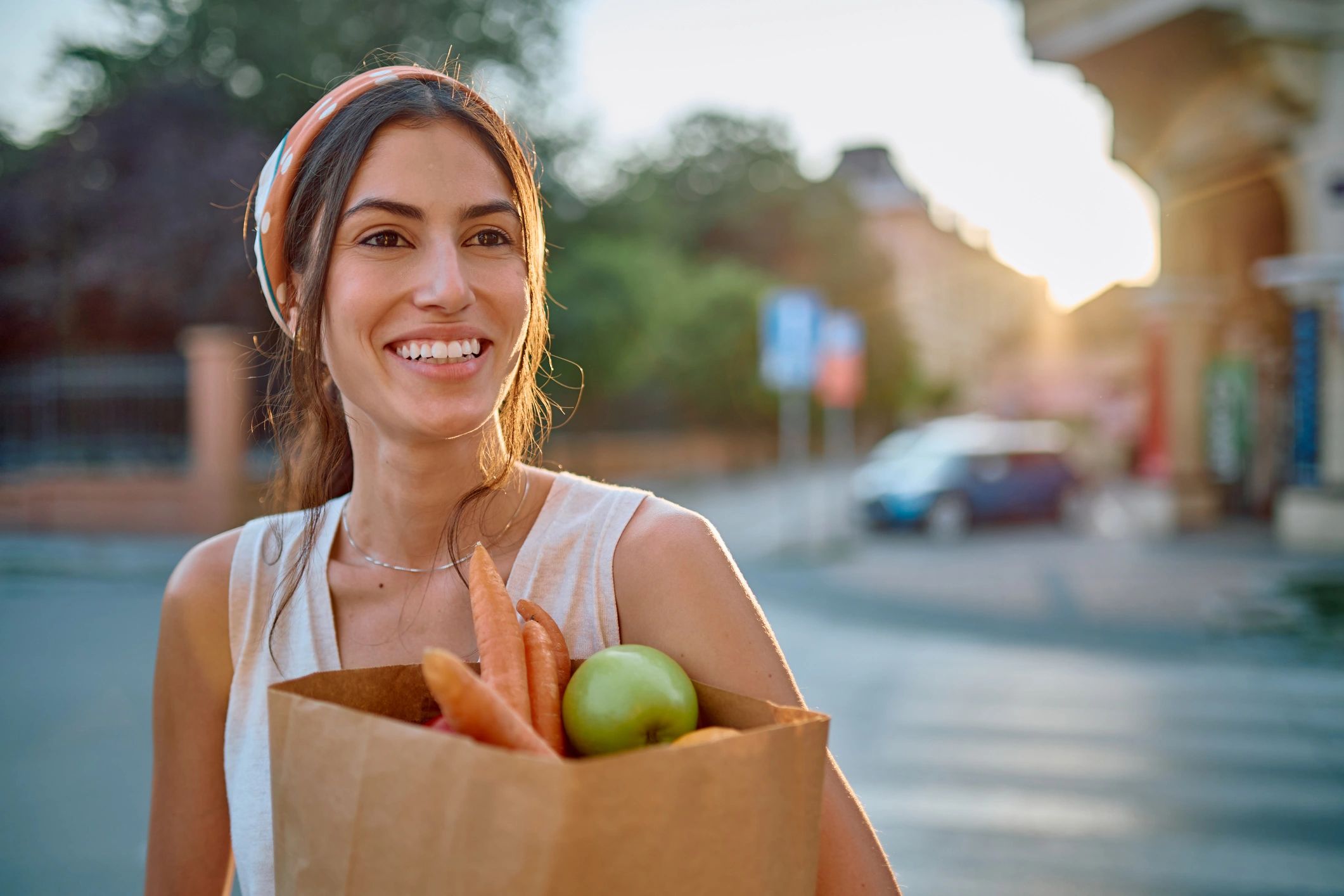 Smiling person carrying groceries in a paper bag at sunset